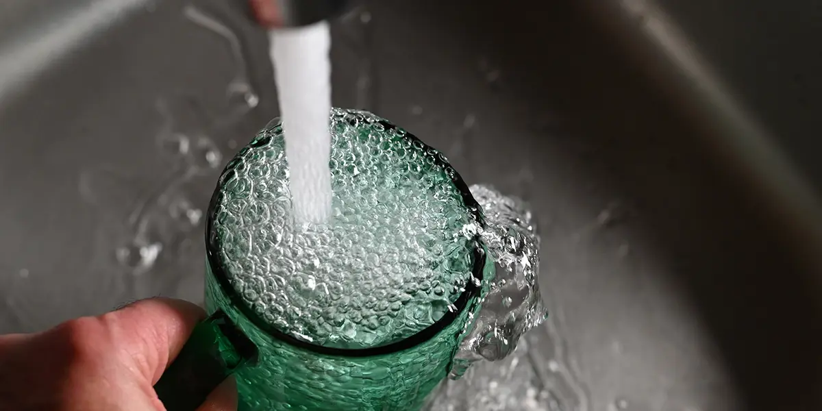 Nahaufnahme eines grünen Glases, das unter einem Wasserstrahl in einer Spüle gefüllt wird, wobei das Wasser überläuft.