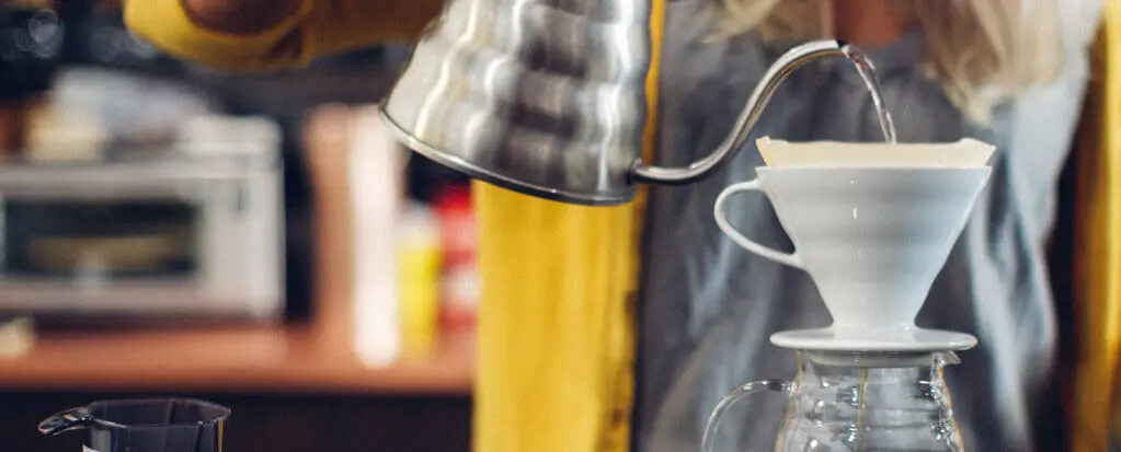 Person gießt Wasser aus einer Metallkanne in einen Handfilter über einer Glaskaraffe zur Kaffeezubereitung.