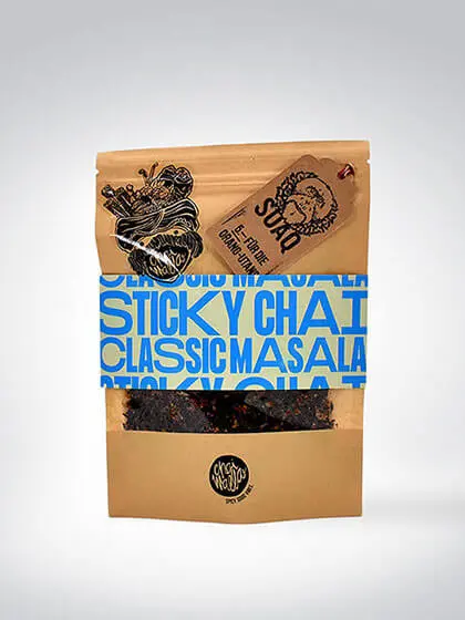 Eine braune Verpackung mit blauen Buchstaben, die Sticky Chai Classic Masala Tee beschreibt. Die Verpackung zeigt künstlerische Illustrationen und das Logo der Marke Cha Cha Chai.